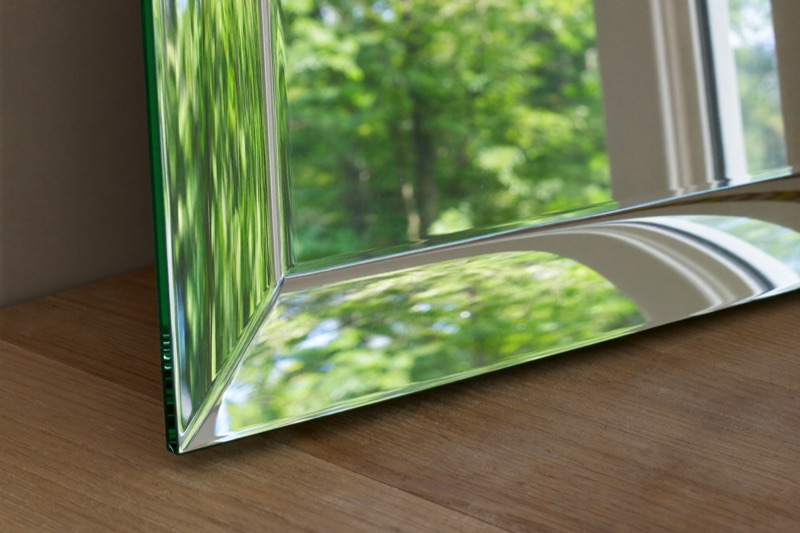 Close view of a polished beveled mirror edge on an oak bench, reflecting summer trees.