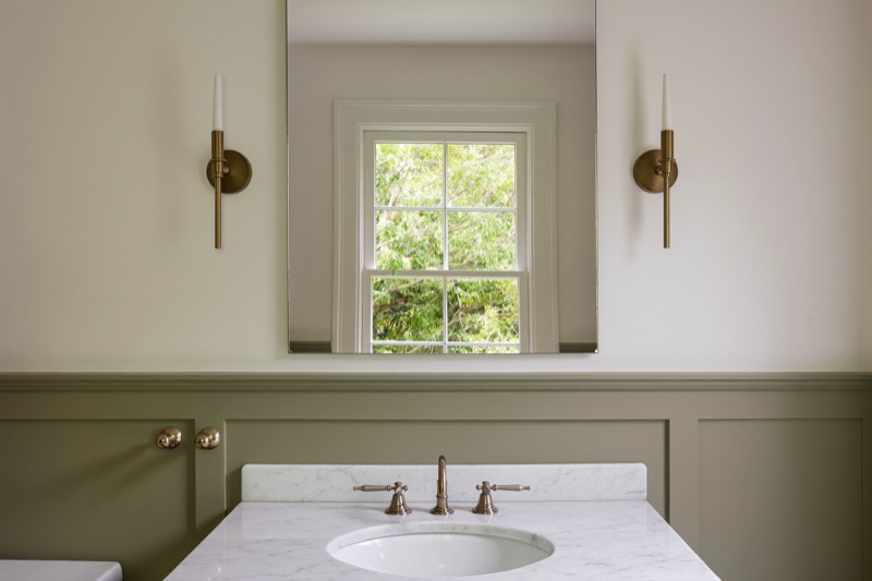 A frameless mirror above a marble vanity in a powder room with sage wainscoting.