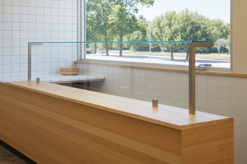 A clear glass sneeze guard on stainless posts above a white oak counter.