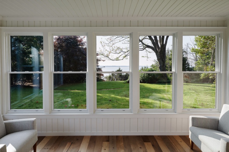 A sunroom wall of white double-hung windows with clear insulated glass, looking onto a lawn and bay.