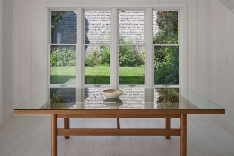 A clear glass top on an oak dining table in front of tall white windows.