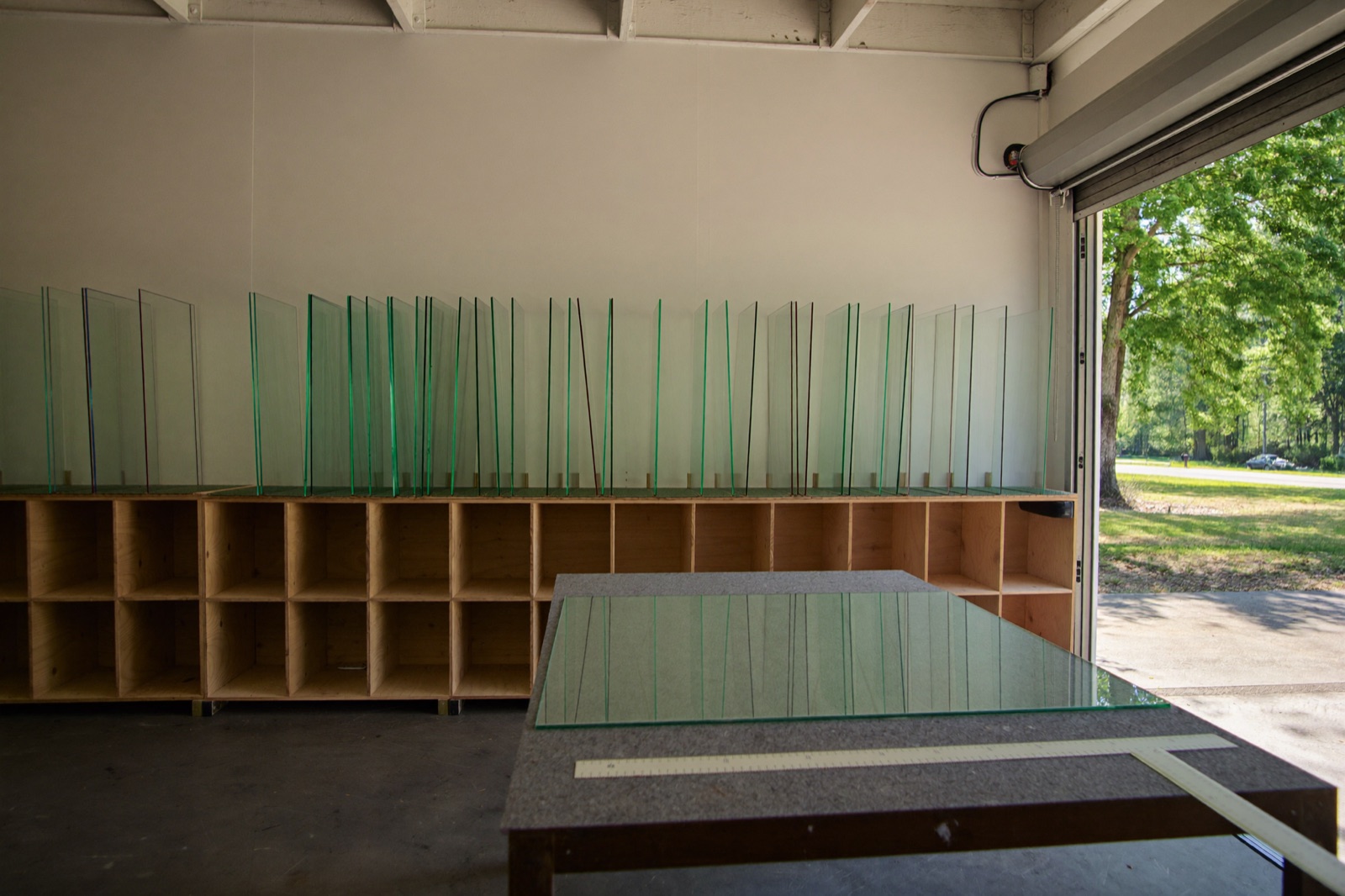 Clear glass sheets standing on edge on a plywood rack, with a felt cutting table and an open door onto summer trees. Illustrative.