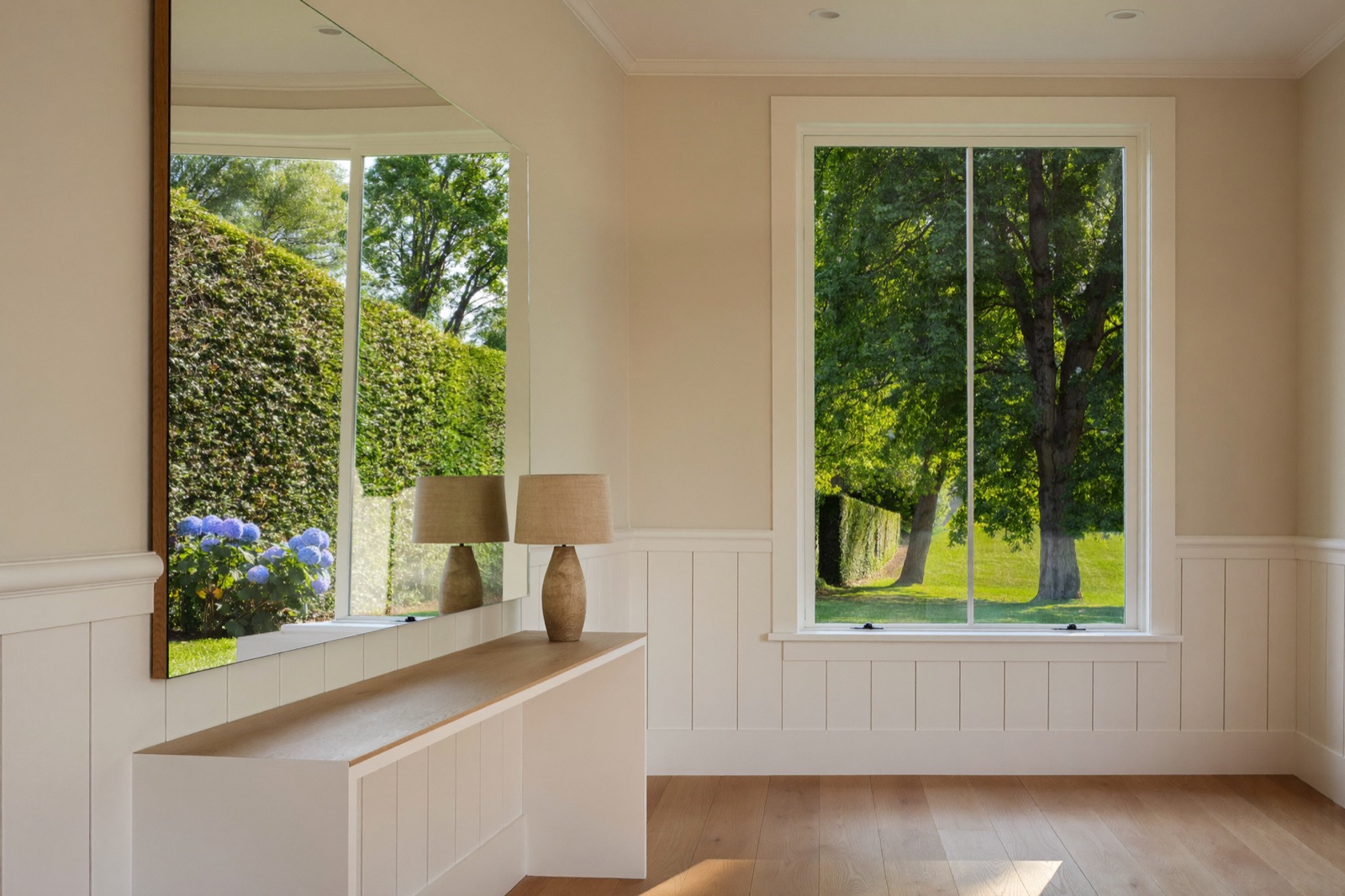 A large frameless wall mirror above a white oak console, reflecting a hedge and blue hydrangeas, in a white wainscoted hall. Illustrative.