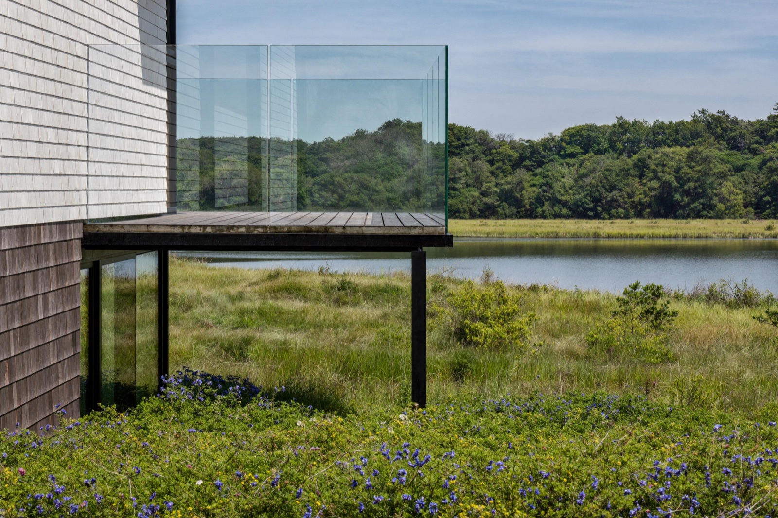 A frameless clear glass deck railing on a shingle house above marsh grass and a bay. Illustrative.