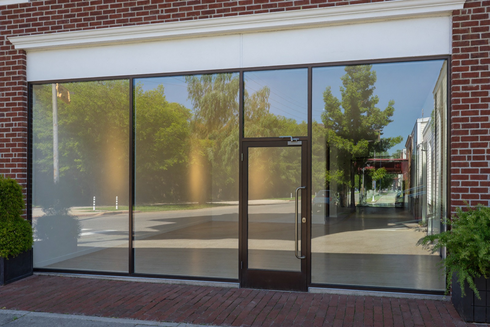 A red brick storefront with tall clear glass in slim bronze frames and a glass entry door, reflecting summer trees. Illustrative.